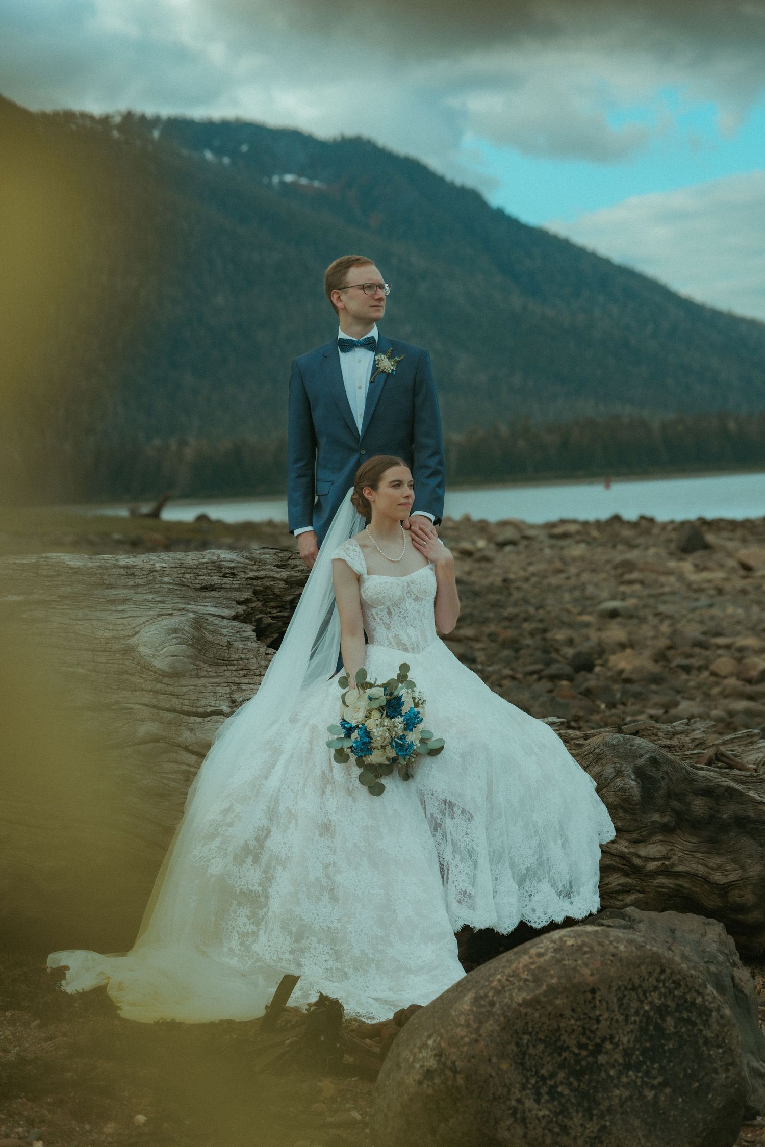 Bride and groom pose on rocky and rugged Alaska beach with mountain views on their wedding day.