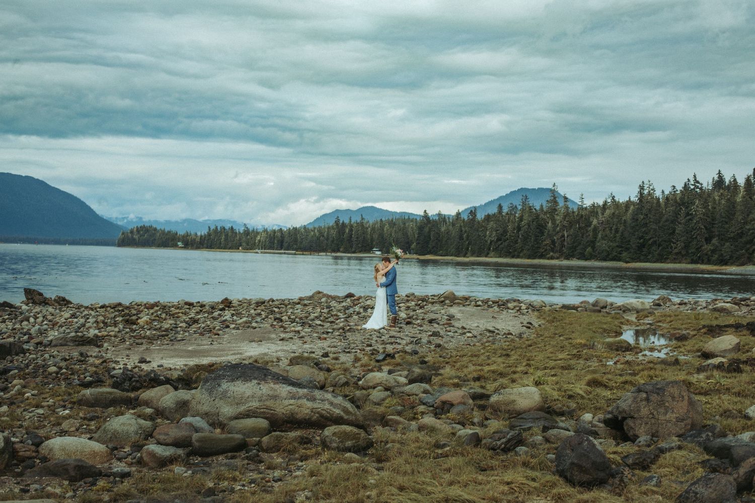Bride and groom embracing on a rocky shoreline with mountains and evergreen forest backdrop.