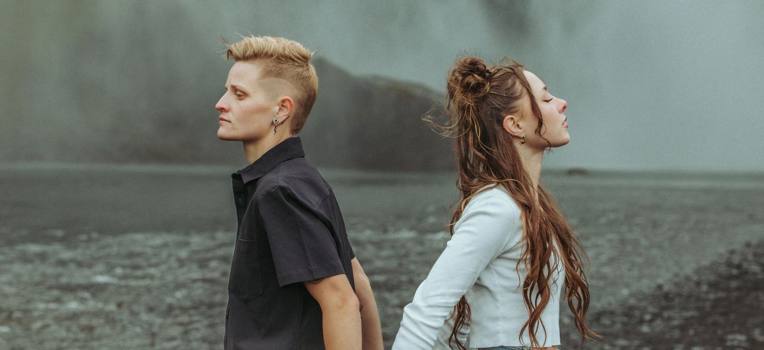 Stylish couple standing back to back on dark volcanic beach near dramatic waterfall in Iceland.