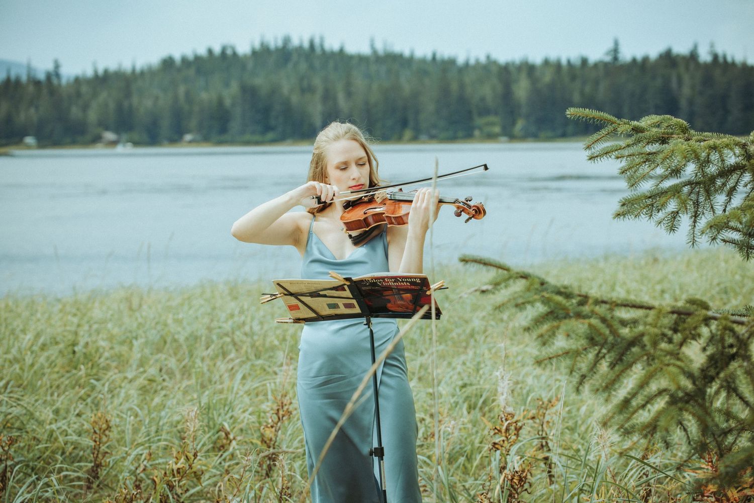 Young musician plays violin outdoors by a tranquil lake surrounded by evergreen forest.