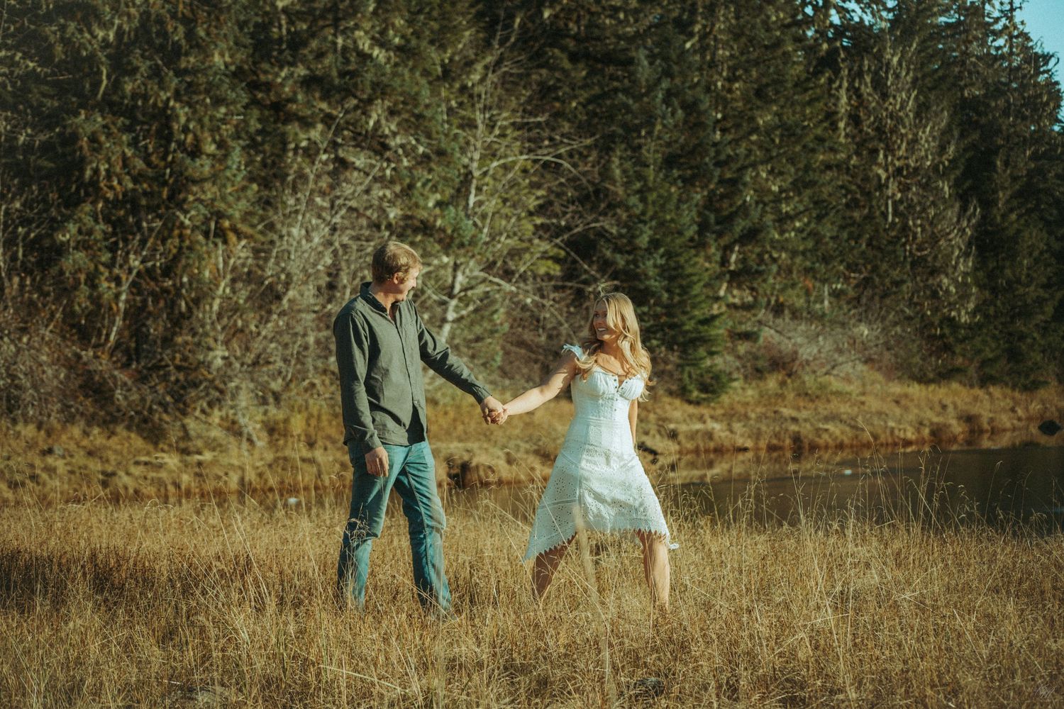 A couple holding hands in a meadow with evergreen trees in the background at golden hour in Southeast Alaska