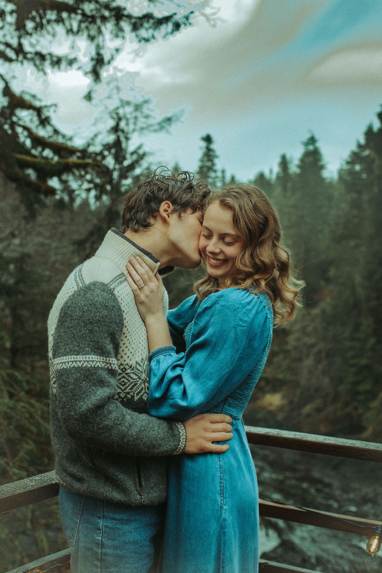 A romantic couple embraces on a deck overlooking a misty evergreen forest, dressed in blue denim dress and grey sweater in Southeast Alaska.
