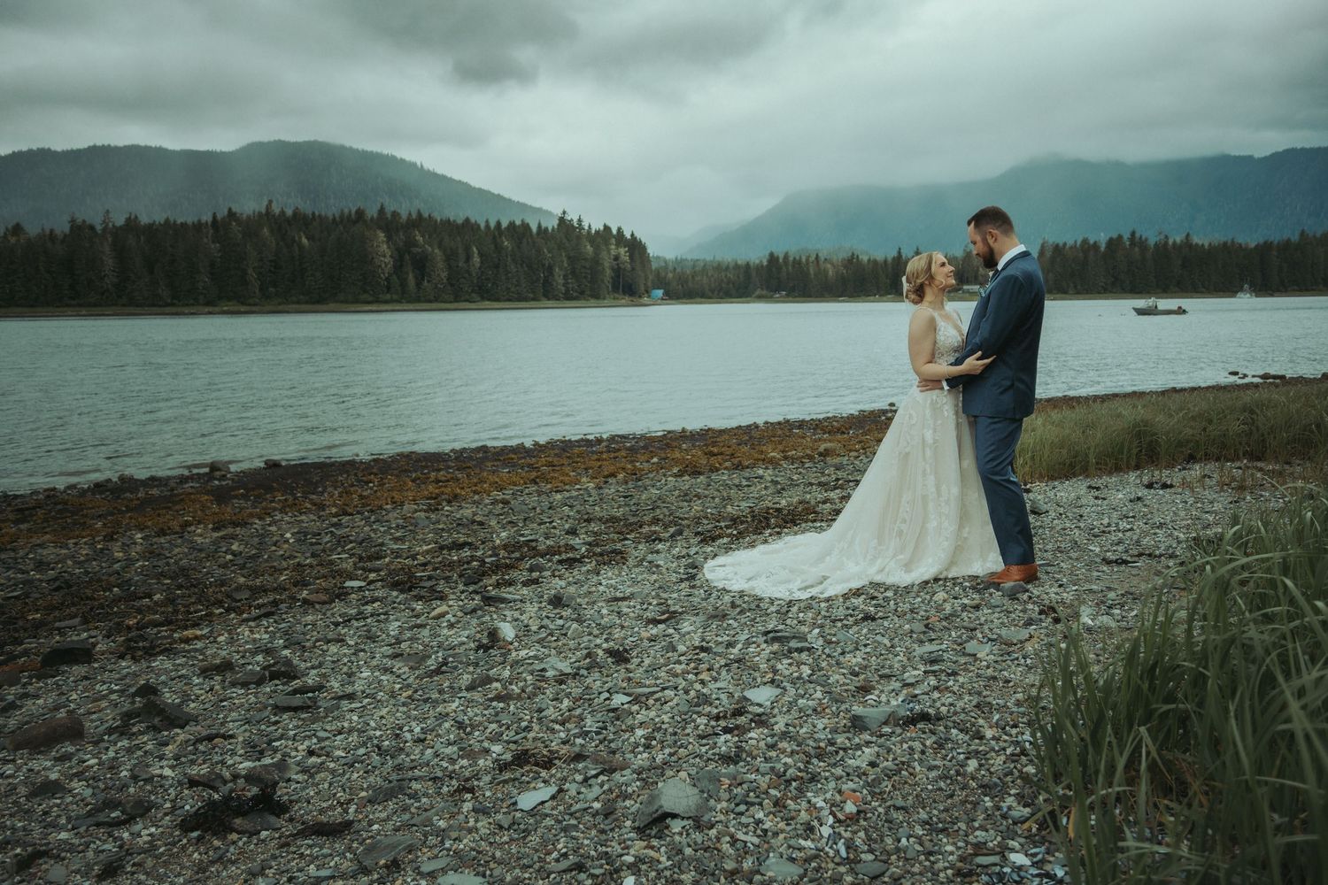 Wedding couple embracing on rocky ocean shore with misty mountain scenery in Alaska.