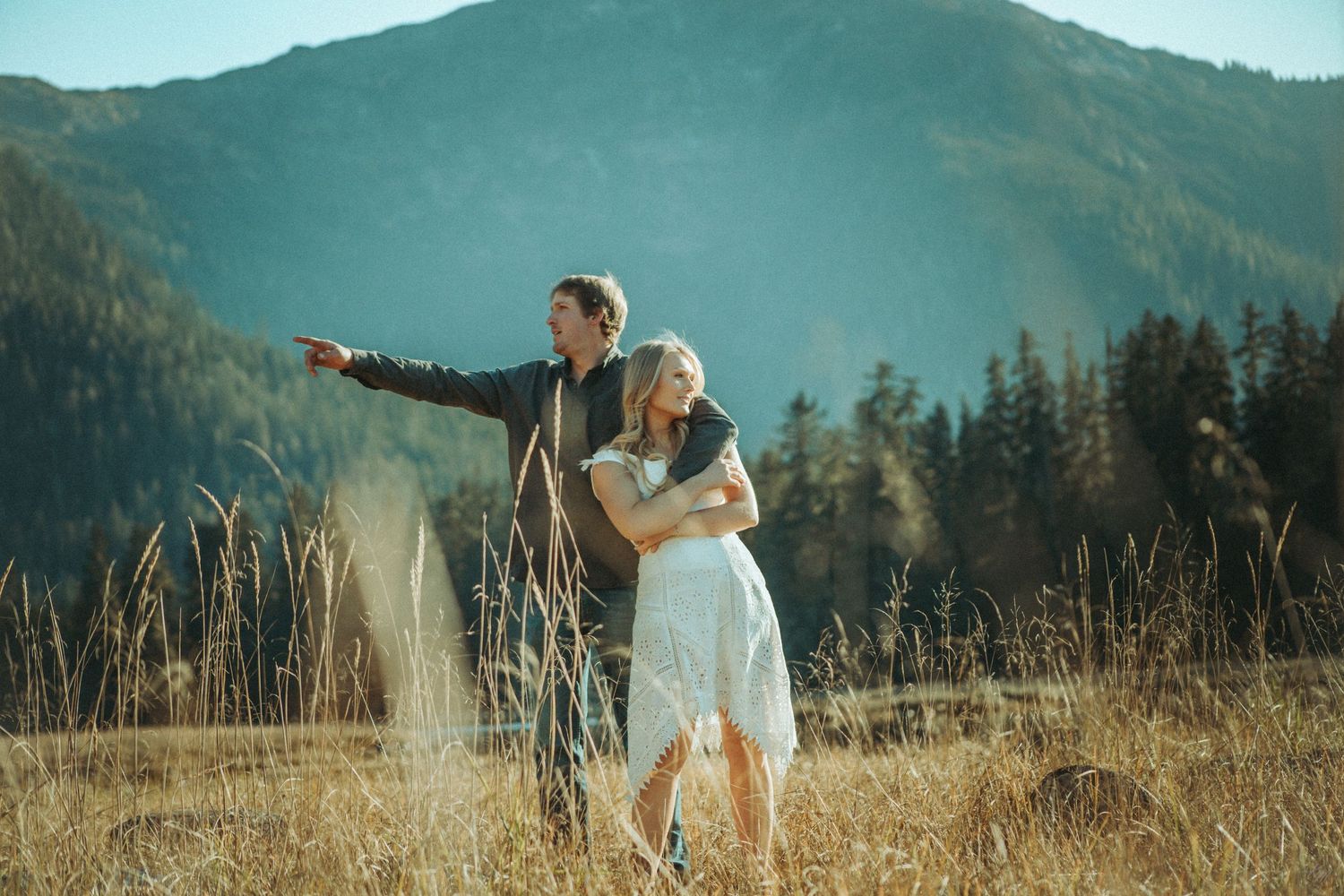 A couple embraces in a golden meadow with majestic mountains and evergreen forest in the background in Southeast Alaska