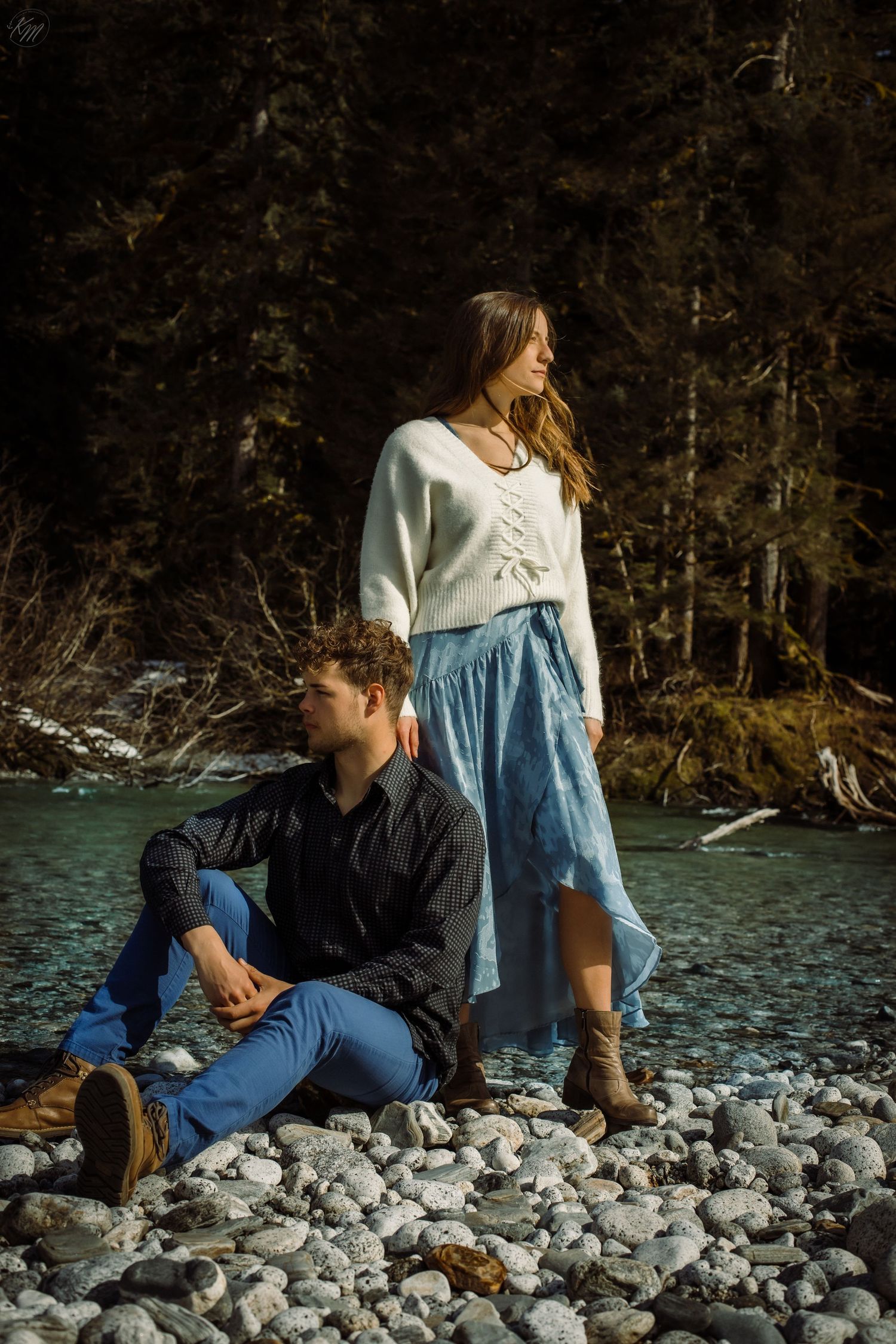Casual couple posing on rocky riverbank with person sitting and other standing in white sweater.