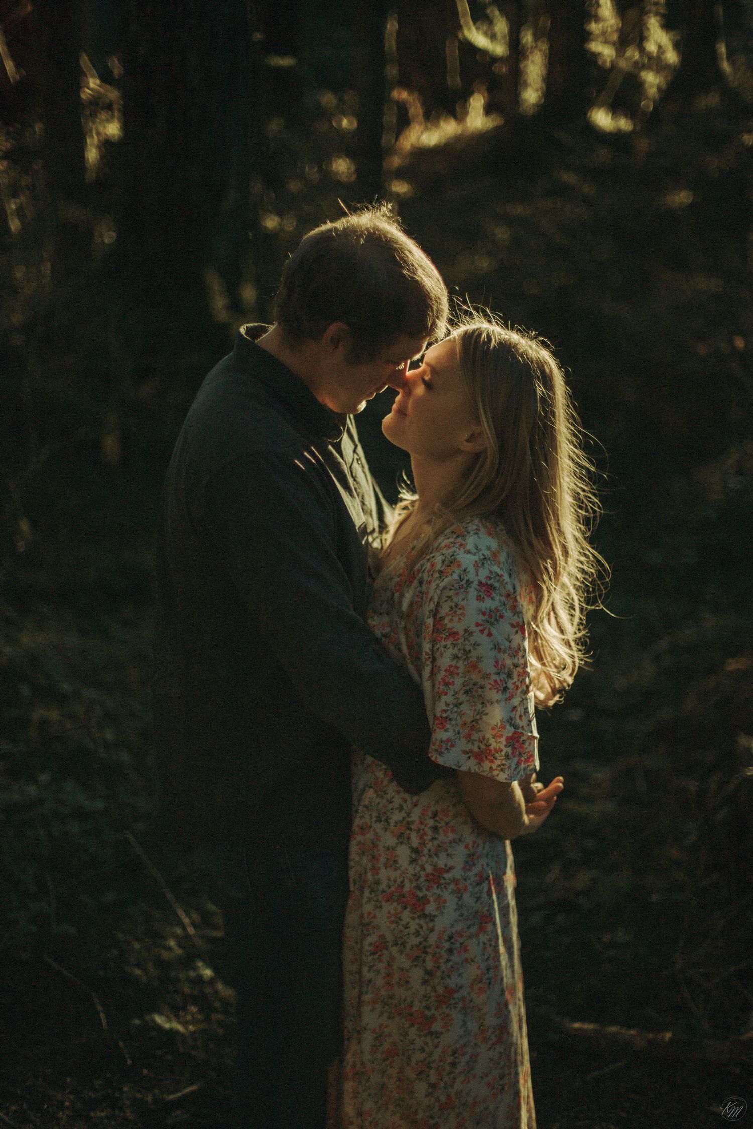 A romantic couple embraces in golden sunlight filtering through trees in Alaska as she wears a floral dress and he wears dark clothing.