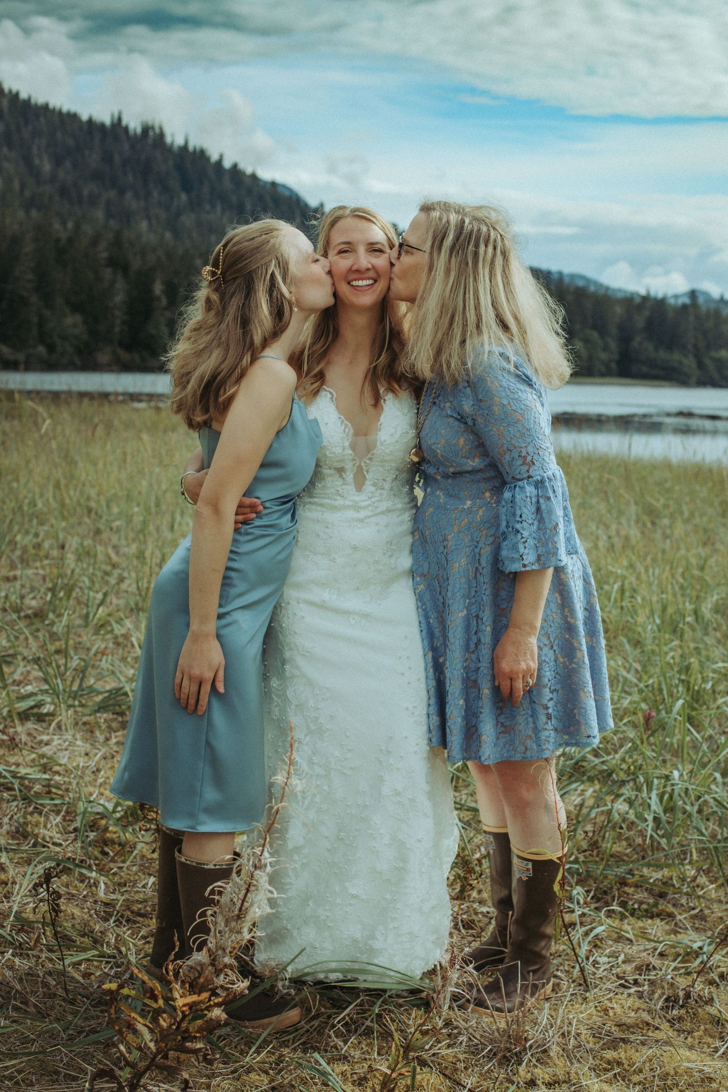 Three women sharing a joyful moment in a field with a lake and mountains in the background.