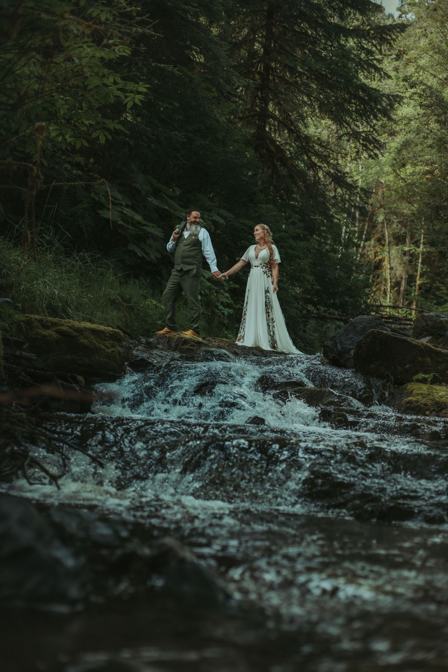 Bride and groom holding hands standing on rocky waterfall in lush green forest wedding photo.