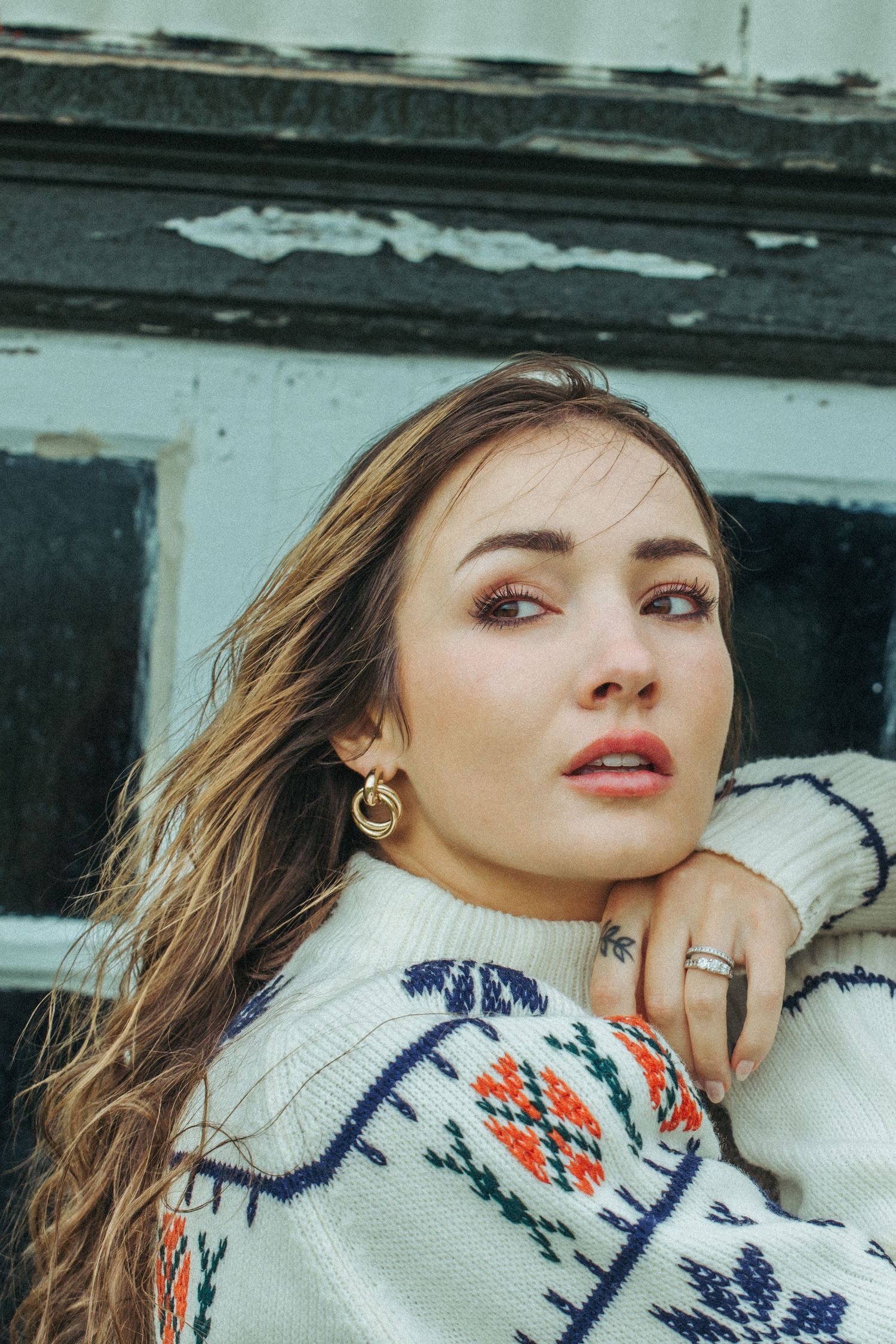 Close-up portrait of a woman in a patterned sweater with windswept hair and jewelry.