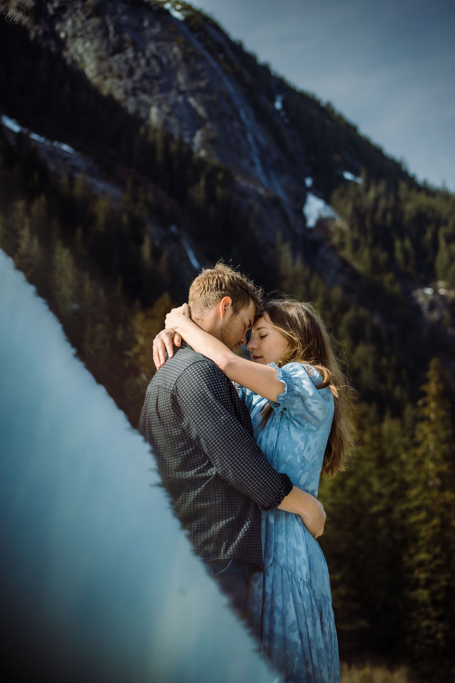 Mountain engagement photos with couple in blue dress and plaid shirt against snowy peaks.