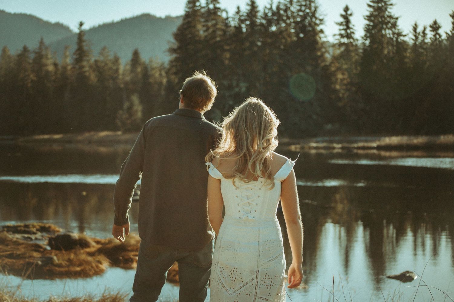 A couple stands together overlooking a peaceful lake during golden hour with mountains and evergreen trees in the background in Southeast AK