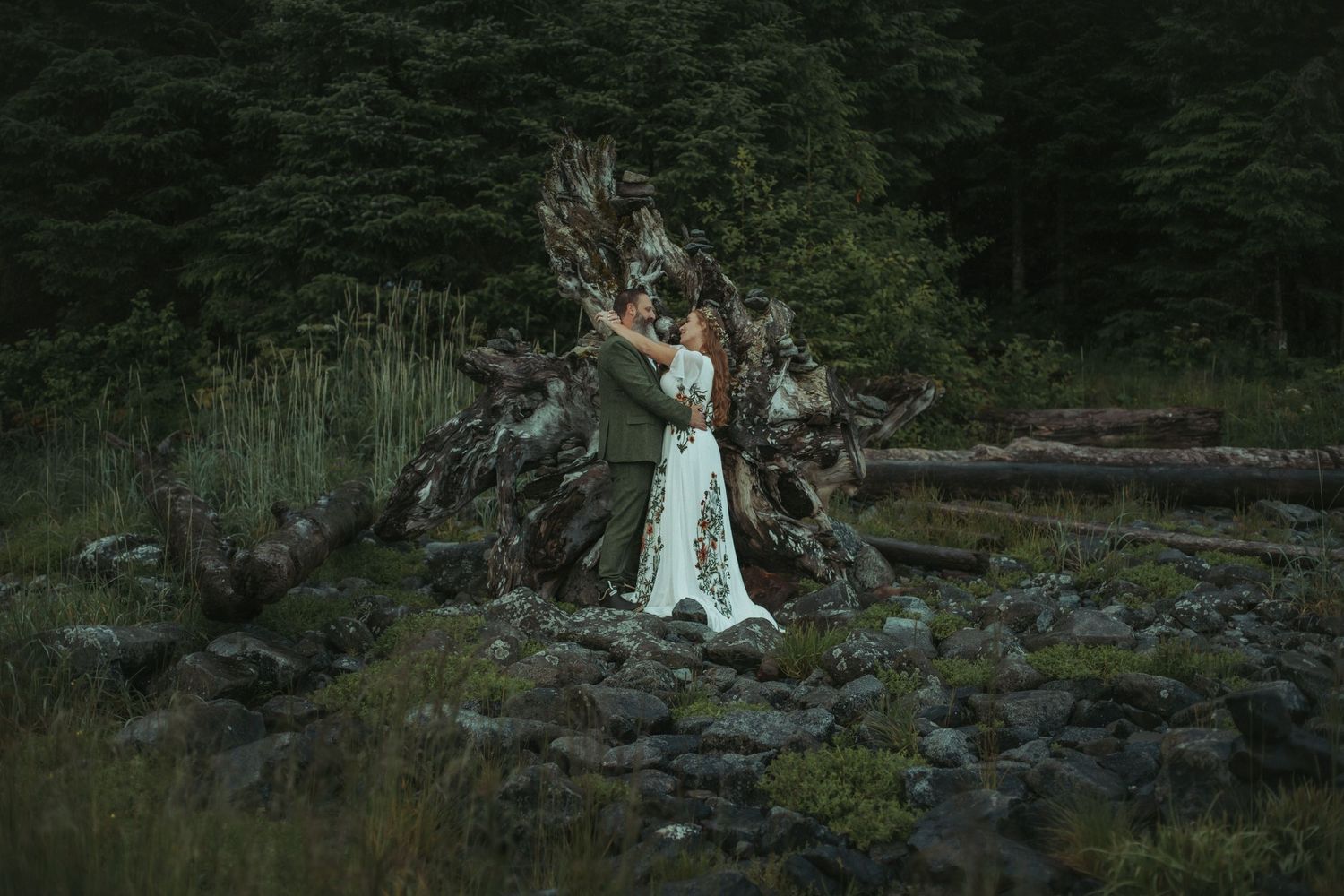 Bride in white lace gown posing beside dramatic driftwood on beach with dark forest behind them in Alaska.