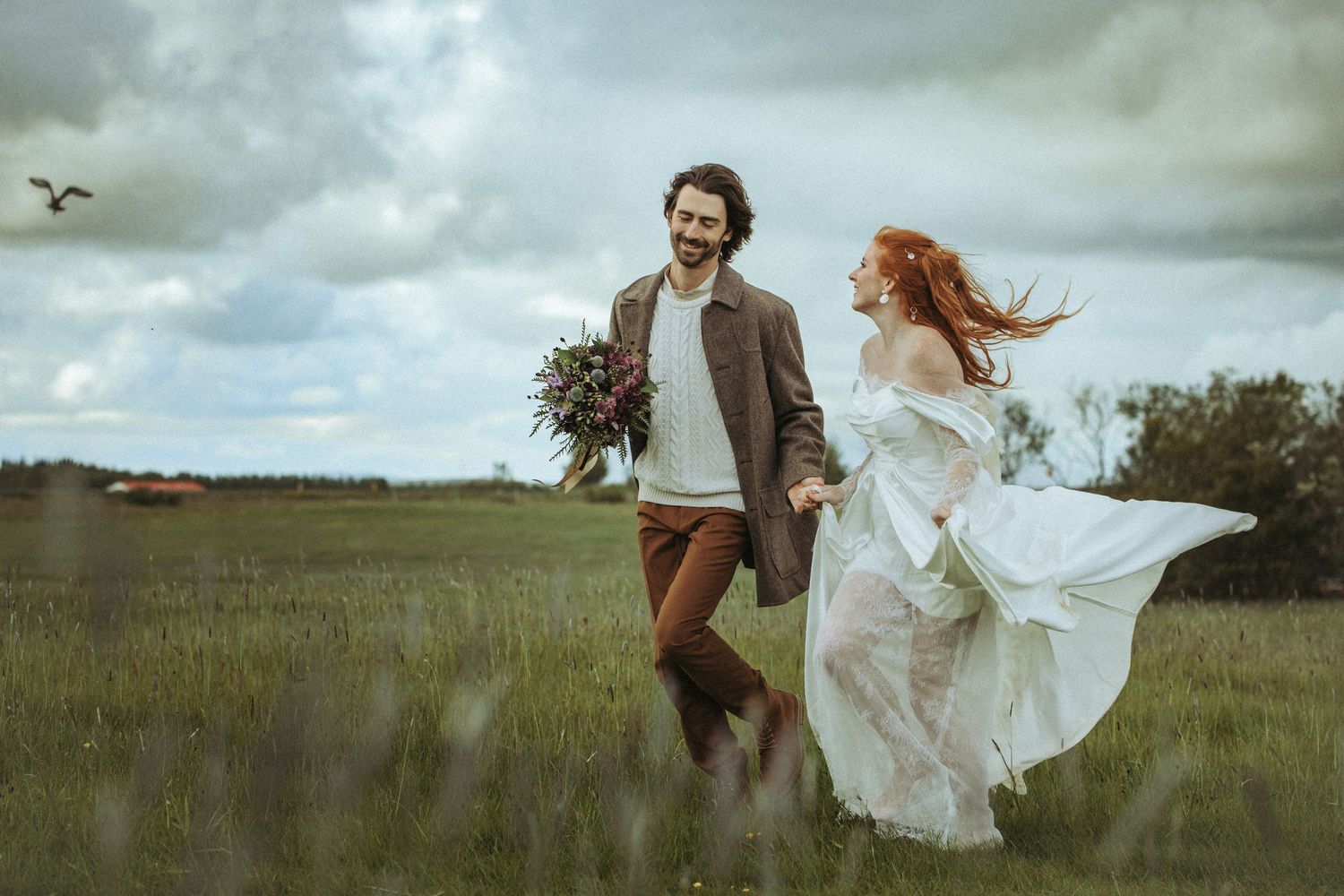 A joyful couple runs through a green field on their wedding day, bride in flowing white dress, groom holding bouquet.