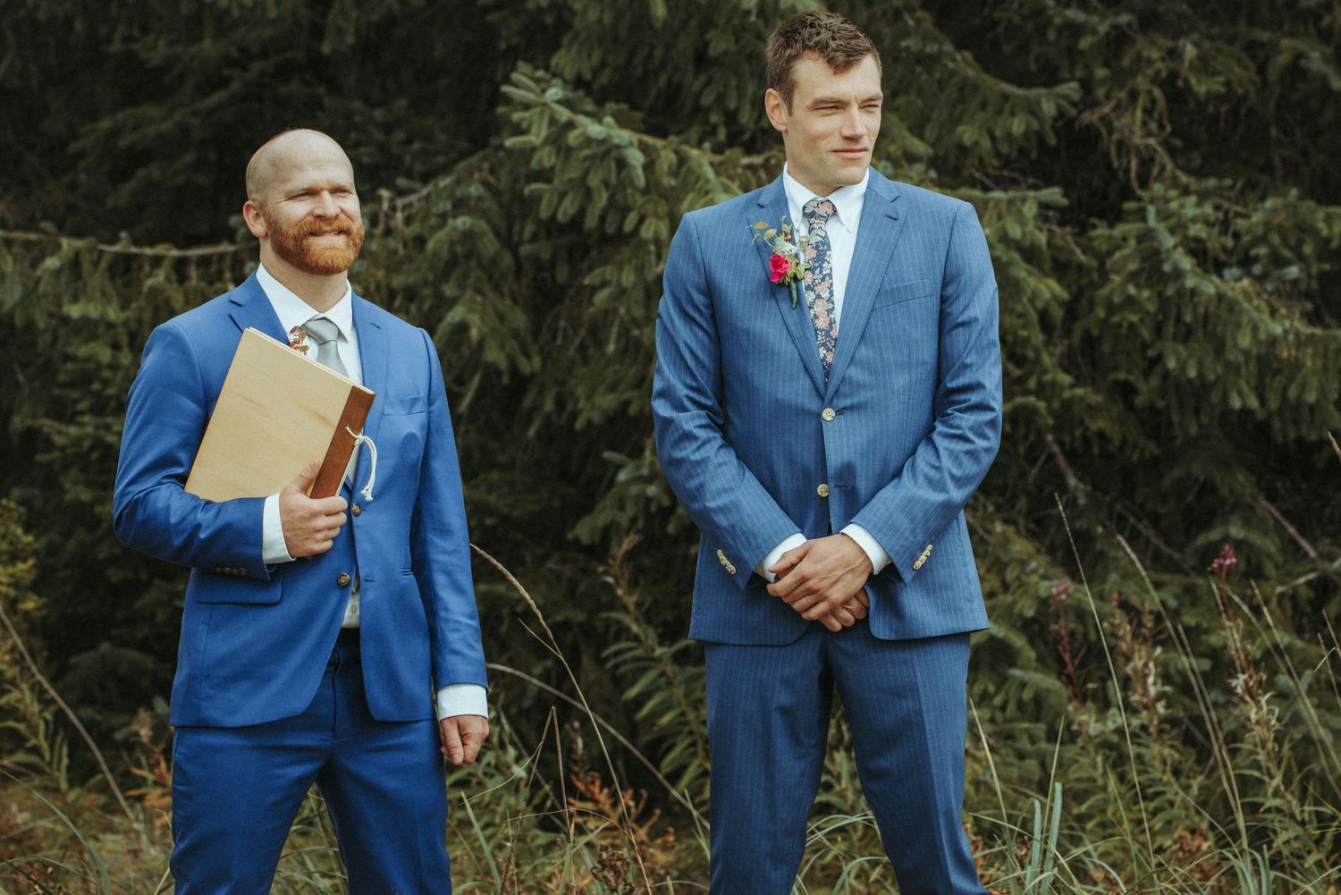 Two men in matching blue suits stand in front of evergreen trees during outdoor ceremony.