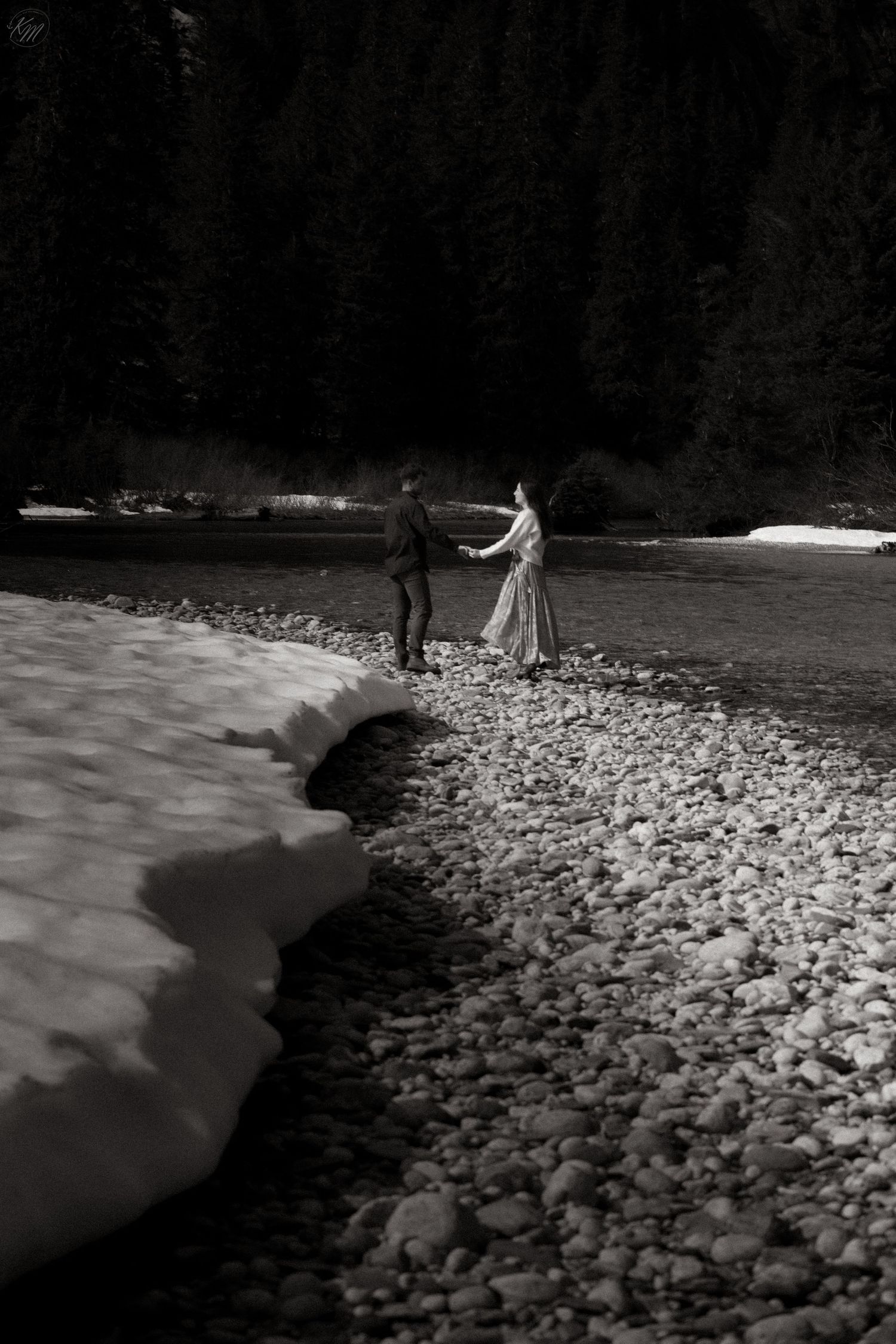 Black and white photo of couple walking hand in hand along snowy riverbank at dusk.