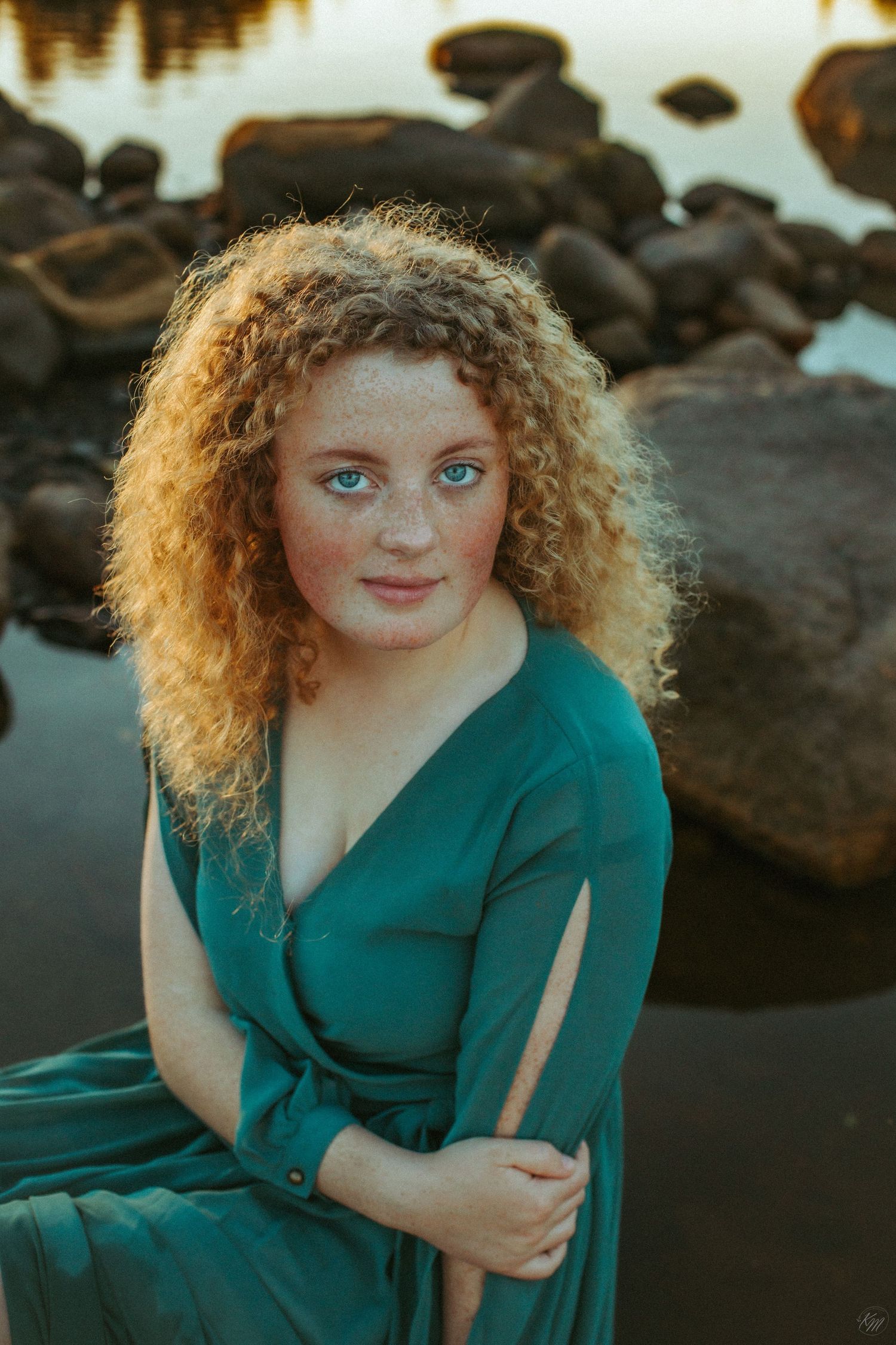 A portrait by the water with curly hair illuminated by golden light and rocks in background.