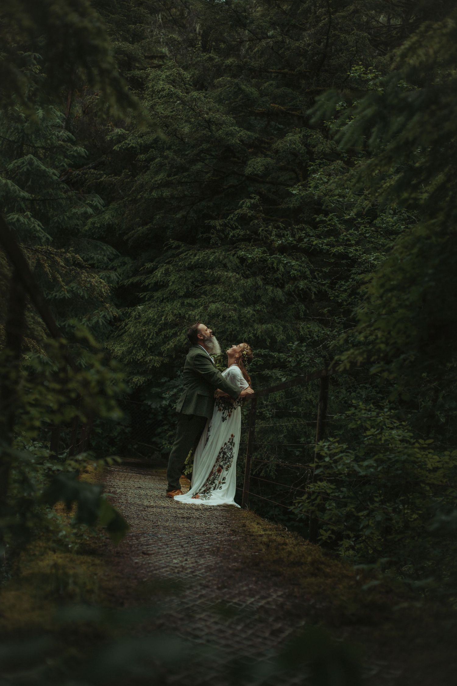 Bride and groom embracing on a forest path surrounded by tall dark evergreen trees.