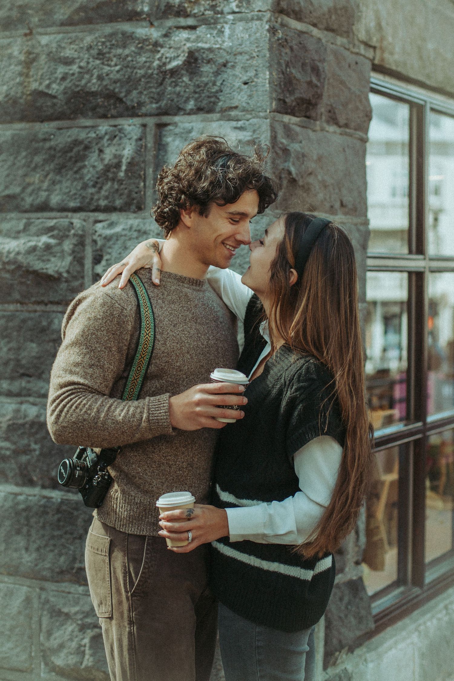 Happy couple sharing coffee and intimate moment together against stone wall exterior with cafe windows.