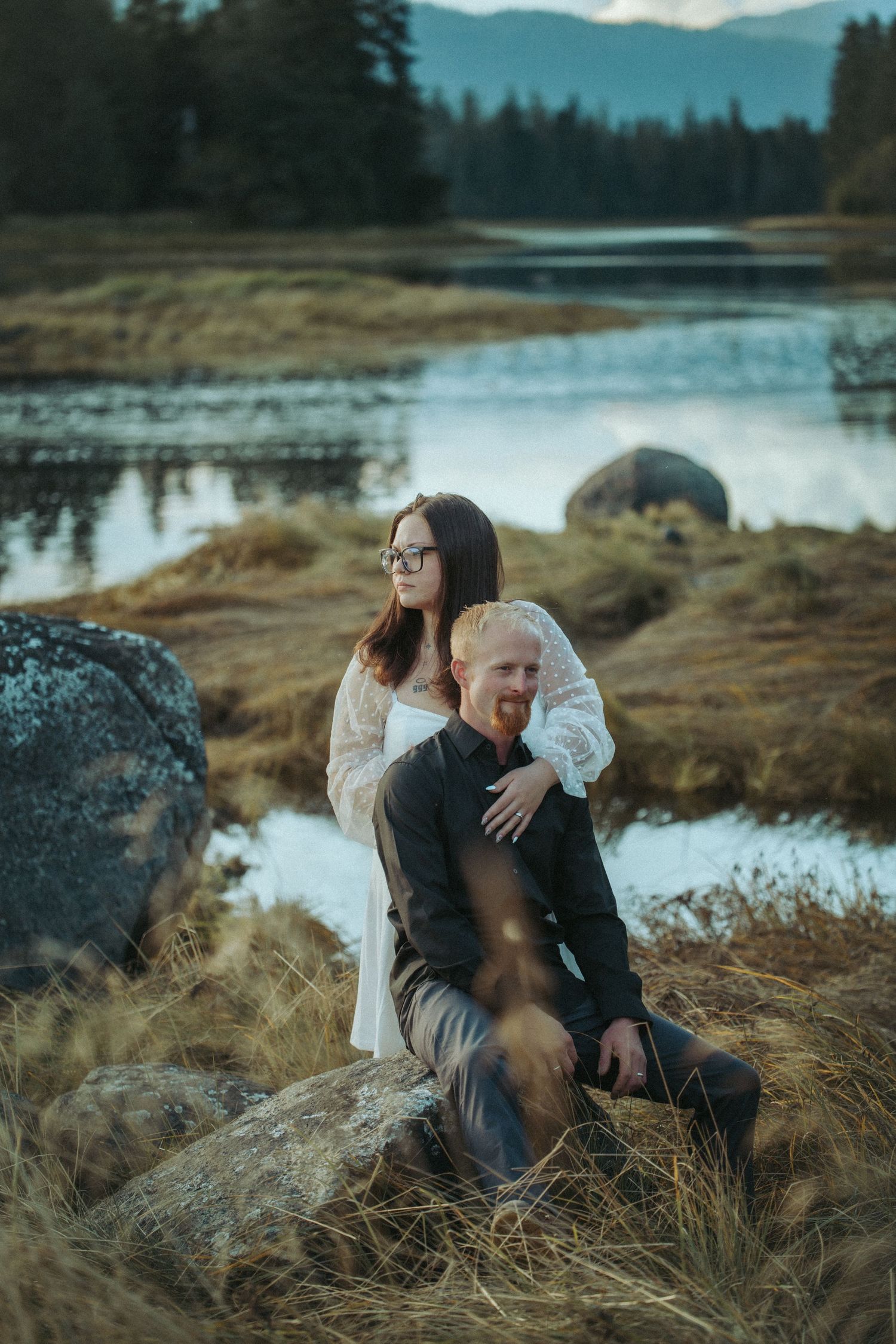 Couple sitting on rocks near a reflective lake surrounded by golden grass and forest in Alaska.