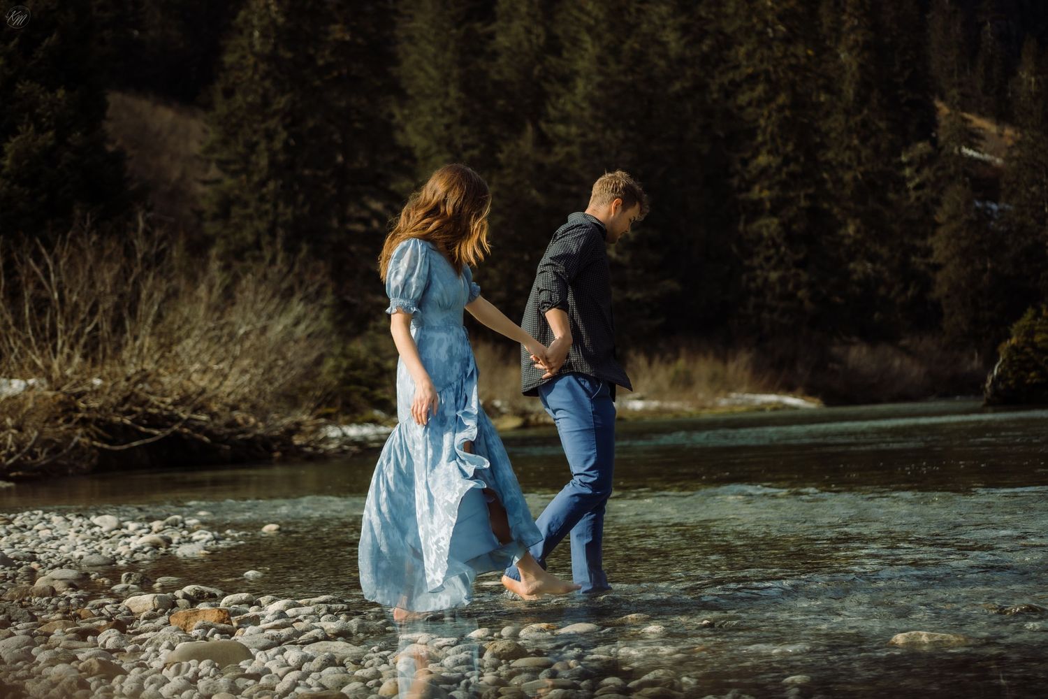 Couple walking along rocky riverbank with snow-capped mountains in background.