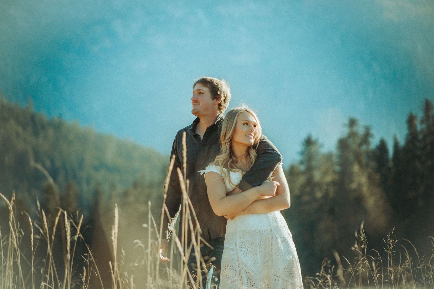 A couple embraces in a golden meadow with majestic mountains and evergreen forest in the background in Southeast Alaska