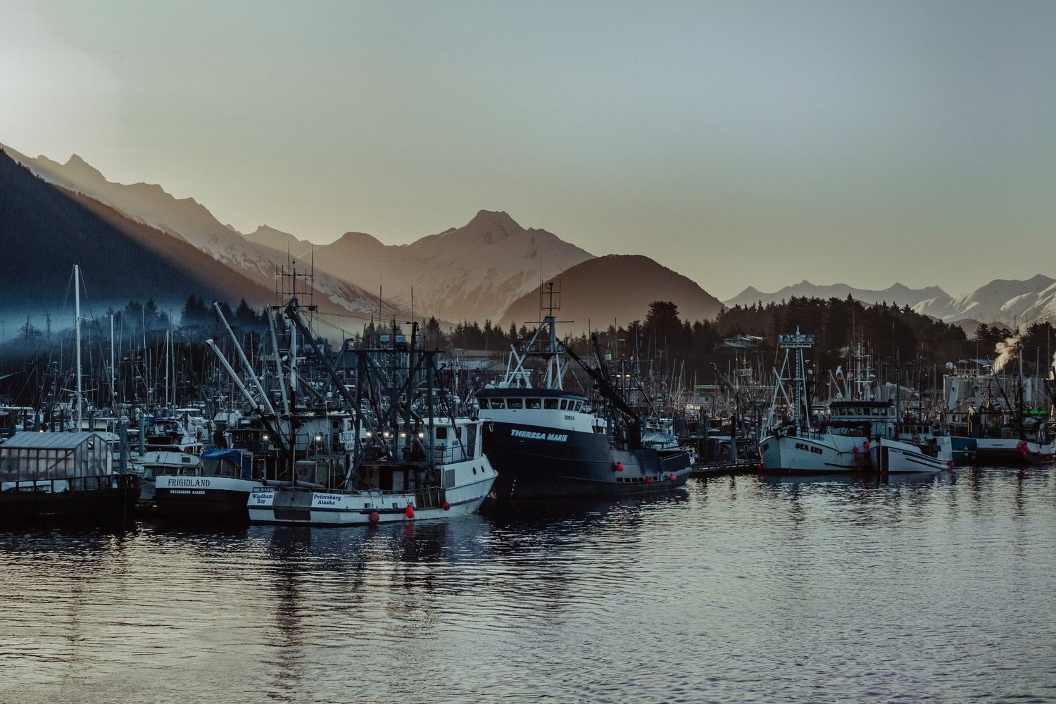 Fishing boats docked in a harbor at dusk with misty mountain peaks in the background.