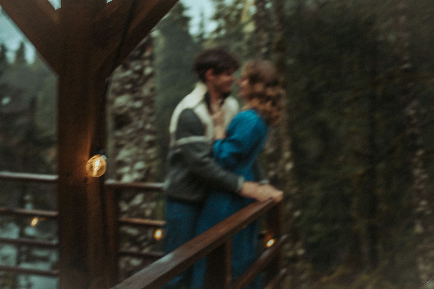 A romantic couple embraces on a wooden deck surrounded by misty evergreen forest, wearing a sweater and denim blue dress in Southeast Alaska