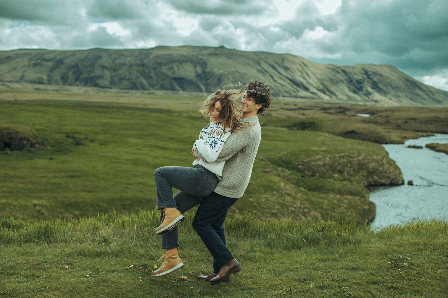 Playful lift pose against backdrop of mountains and meandering river while wearing cozy nordic sweaters for Iceland Engagement.