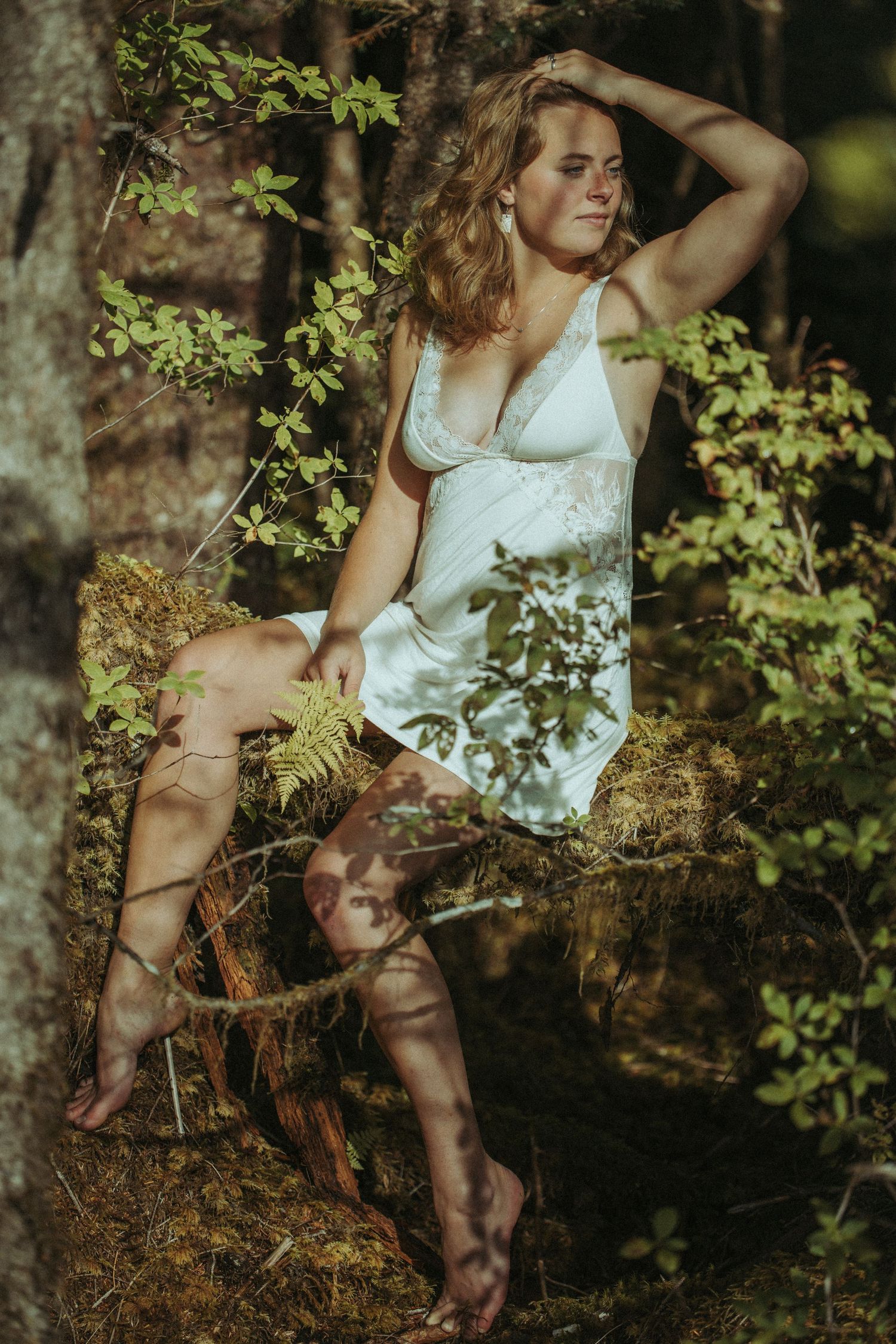 Woman in white slip dress sitting barefoot on mossy log in lush forest setting.