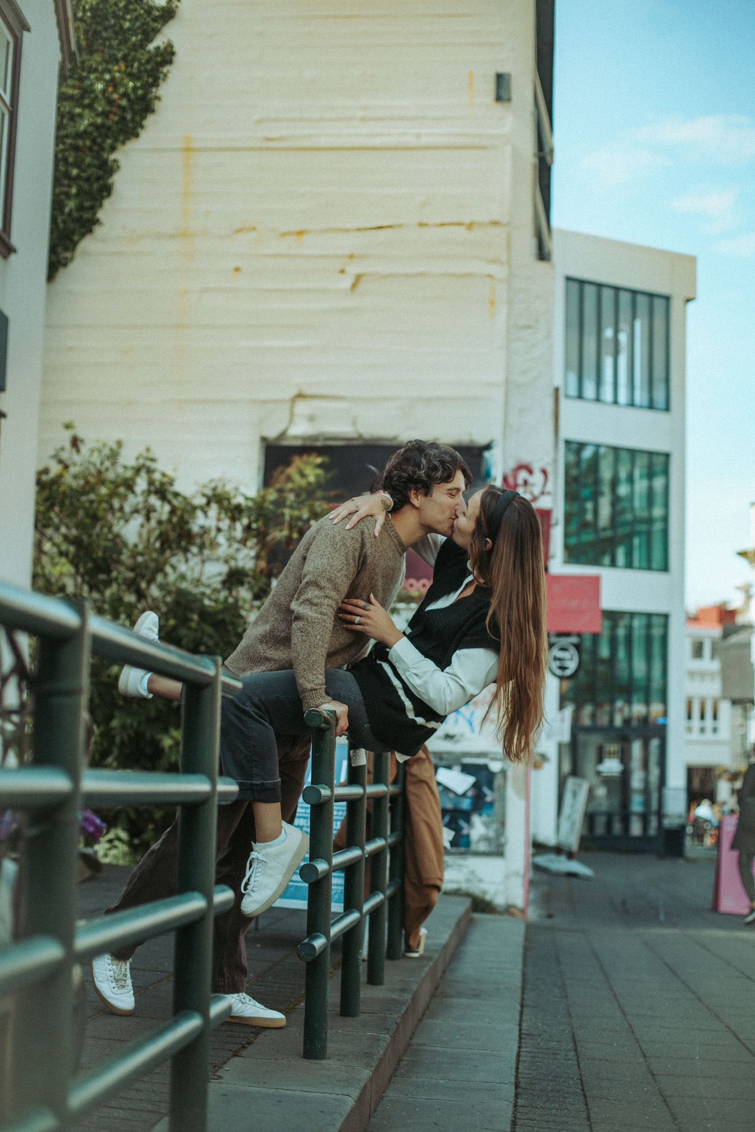 Two people lean against metal railings in a playful urban scene with white buildings during Iceland Engagement session in Reykjavik.