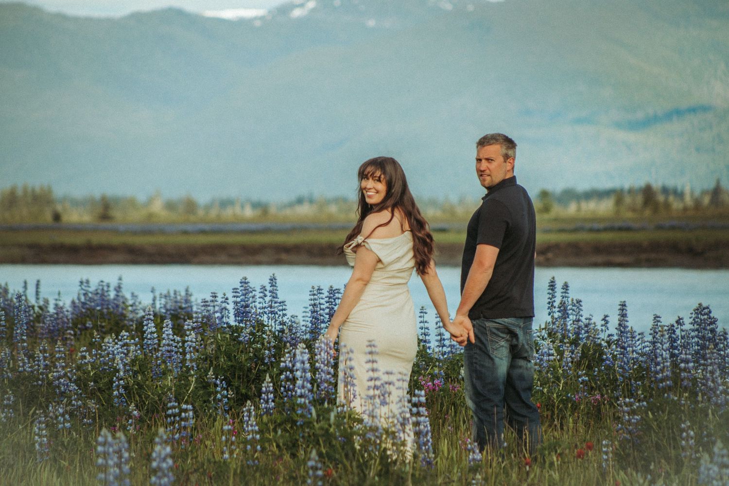 Engaged couple stand in a field of purple lupines by a lake with mountains in the background, Southeast Alaska at golden hour.