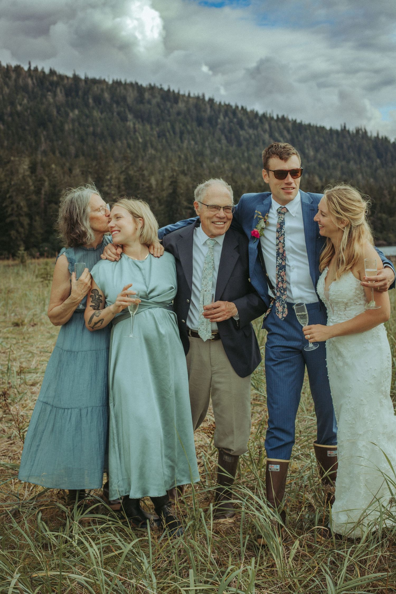 Wedding party in pastel dresses and formal wear stand together in a scenic mountain meadow.