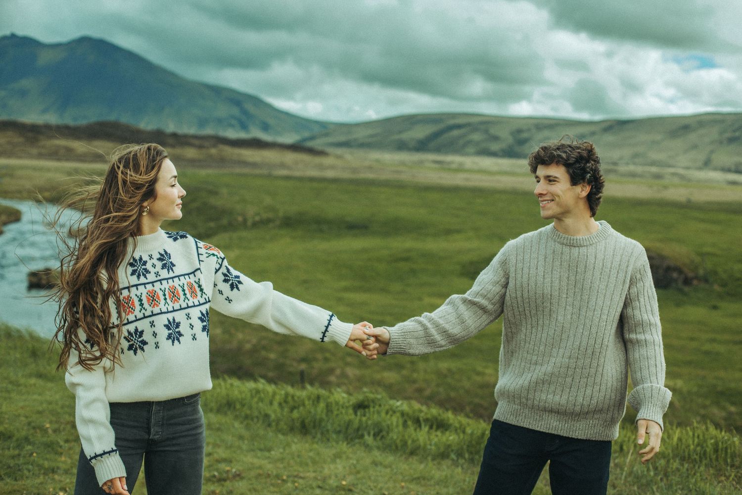 A couple shares a playful moment against a dramatic mountainous landscape with cloudy skies while wearing cozy sweaters.