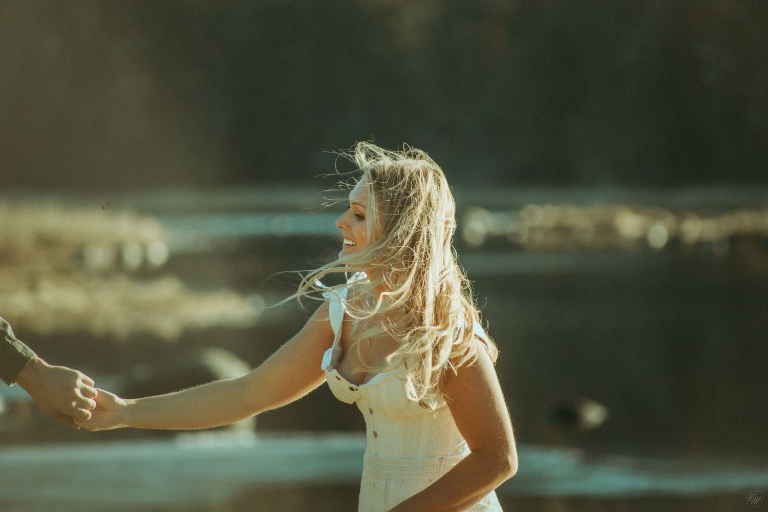 Person in a white dress with flowing blonde hair reaches out during golden hour lighting by a waterfront location in Southeast Alaska.