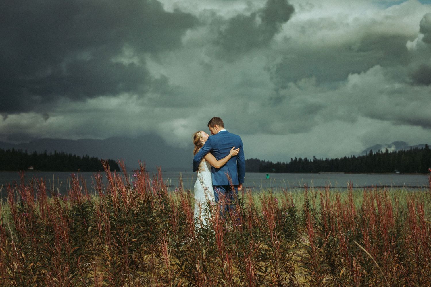 A bride and groom embraces in a field of tall grass under moody storm clouds with the ocean in the background at sunset in Alaska.