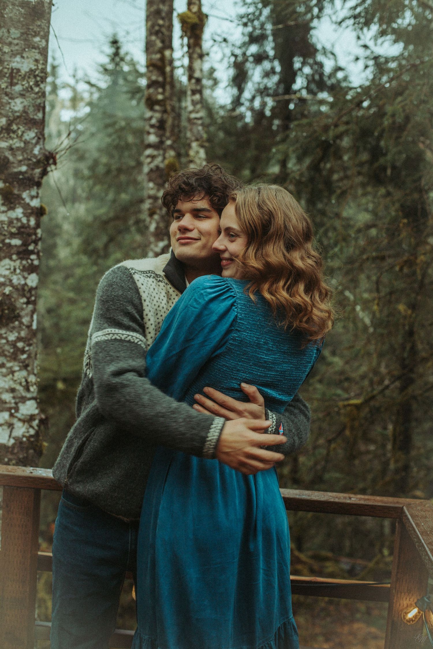 A romantic couple embraces on a wooden deck surrounded by misty evergreen forest, wearing a sweater and denim blue dress in Southeast Alaska
