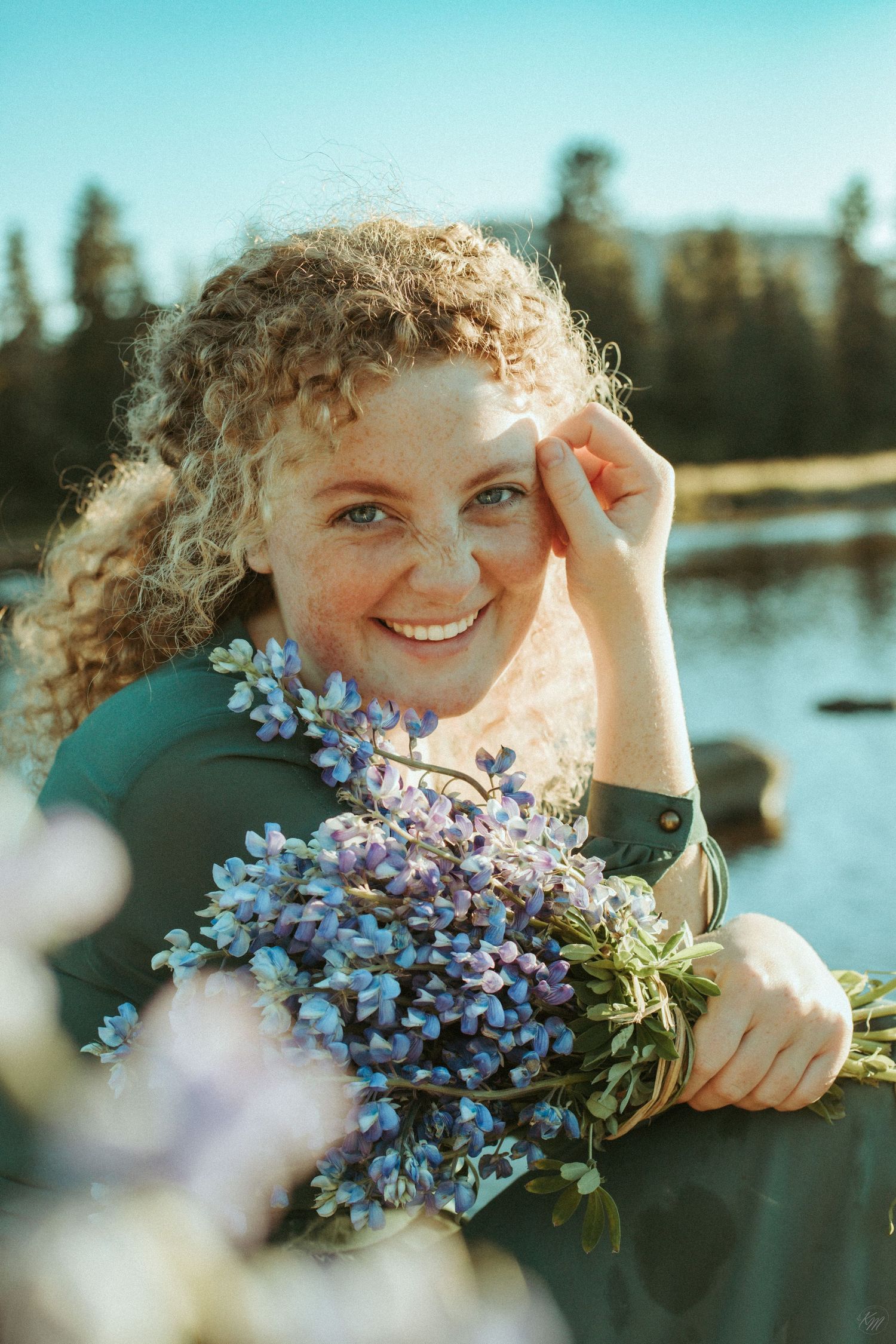 Joyful portrait holding blue wildflowers against mountain lake backdrop.
