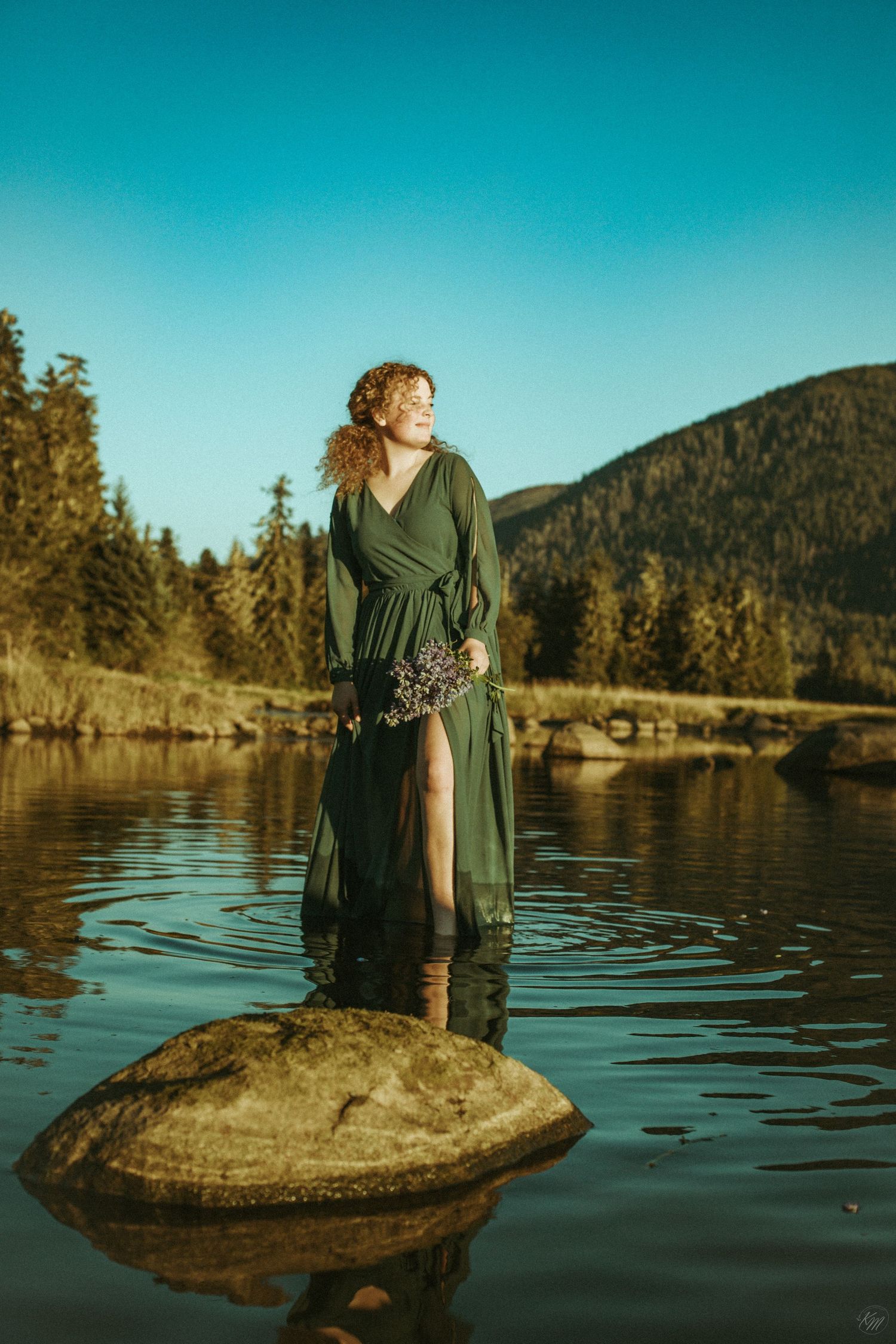 Figure in emerald dress standing on rock in lake with mountains behind.