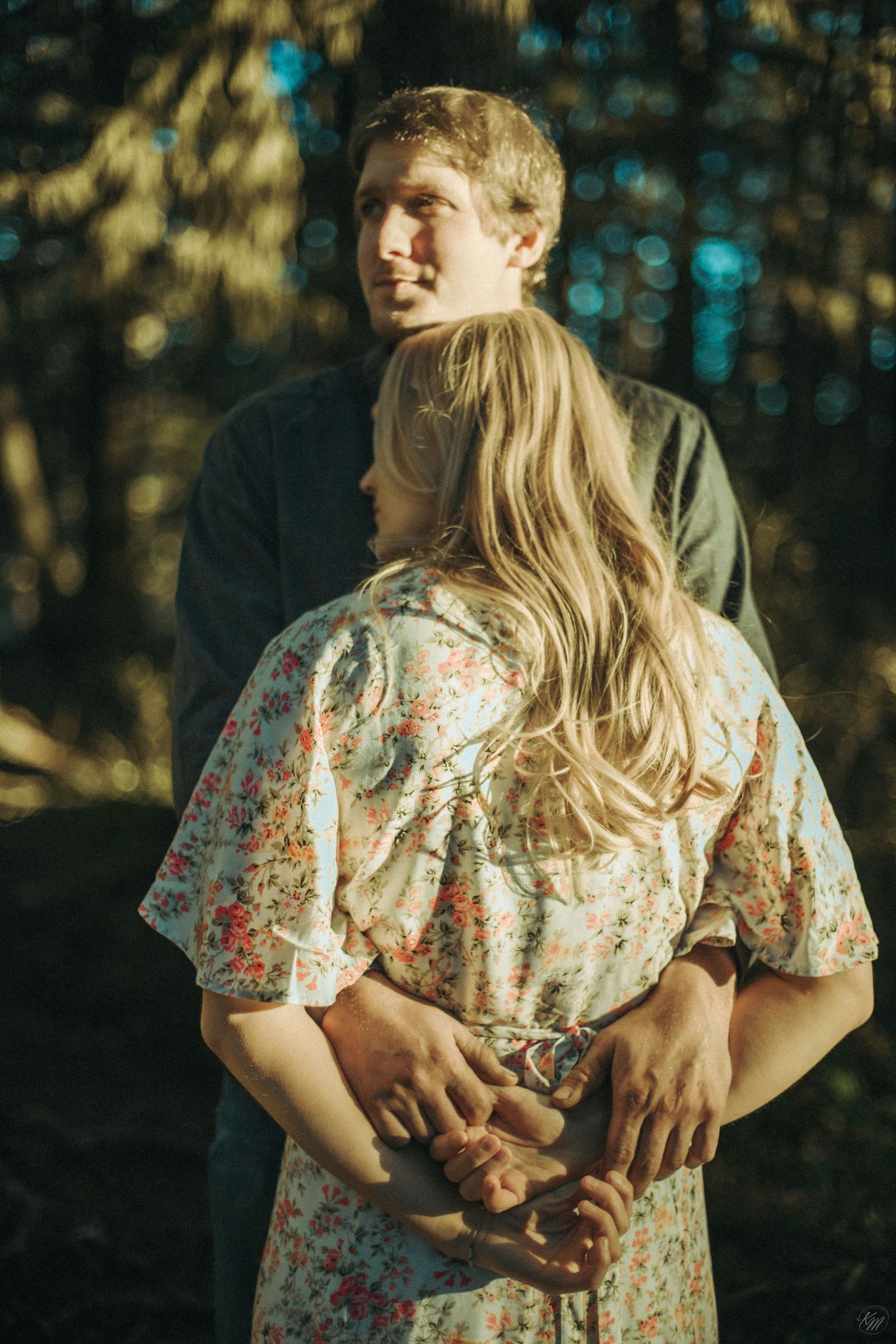 A romantic outdoor portrait shows a couple embracing holding hands in soft golden sunlight, with the woman wearing a floral dress in Alaska