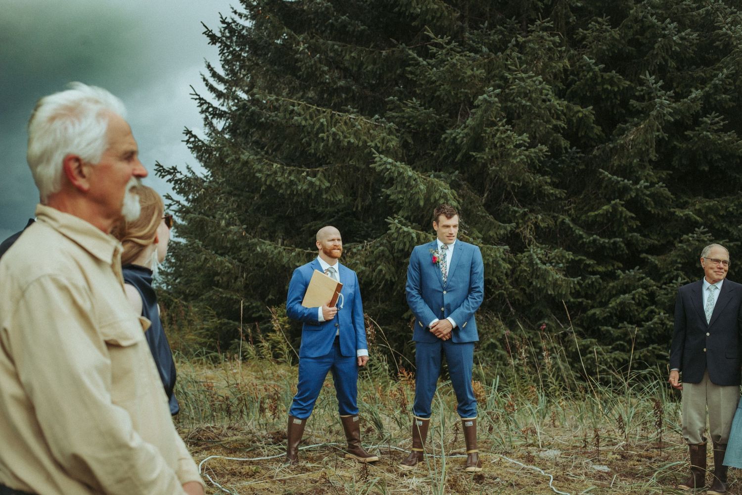 Wedding guests gather on a grassy hillside overlooking a scenic mountain landscape in Alaska.