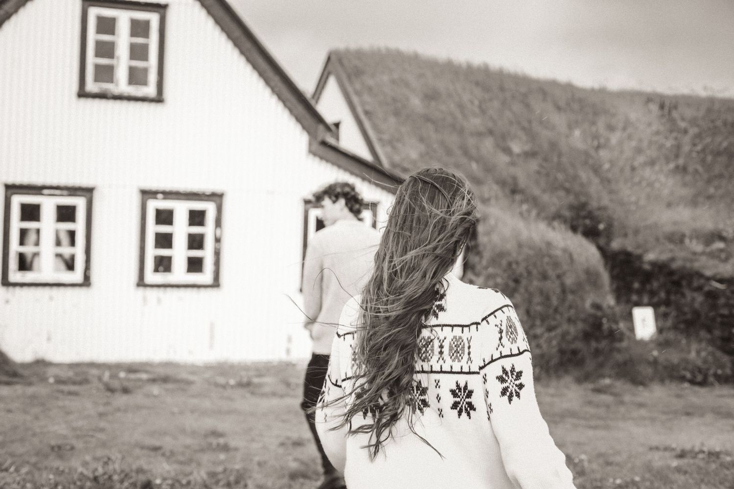 Back view of person in patterned sweater with flowing hair against white Nordic turf house backdrop for Iceland Engagement.