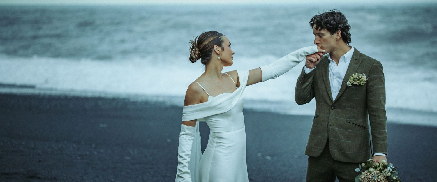 Couple in wedding attire sharing a romantic moment on a black sand beach with dramatic ocean waves.