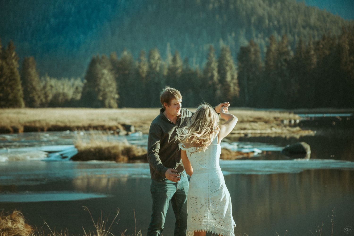 Two people in cute clothing dance joyfully beside a frozen mountain lake surrounded by evergreen forest in Southeast Alaska.