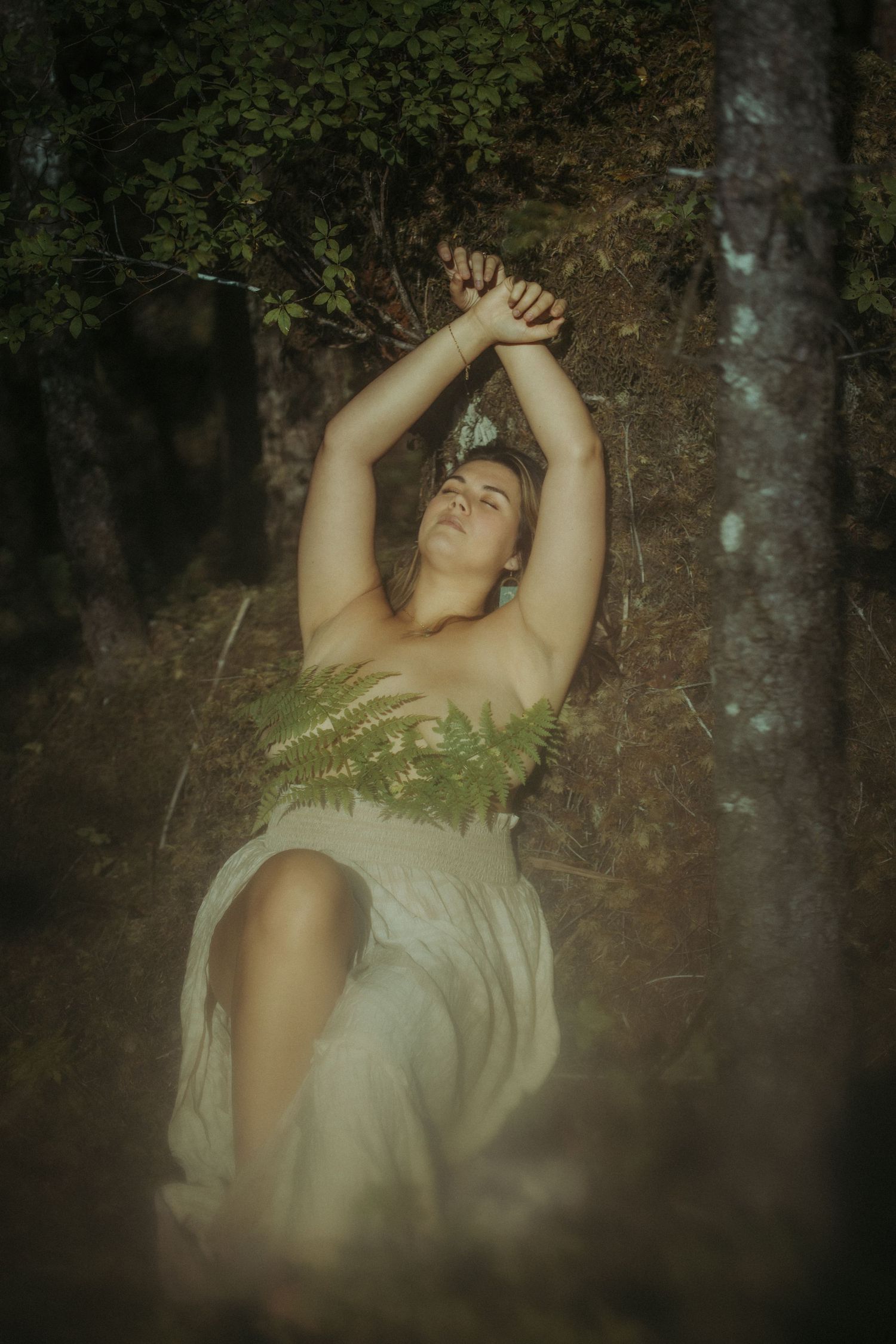Woman in white dress posing dramatically against a tree in a lush green forest setting.