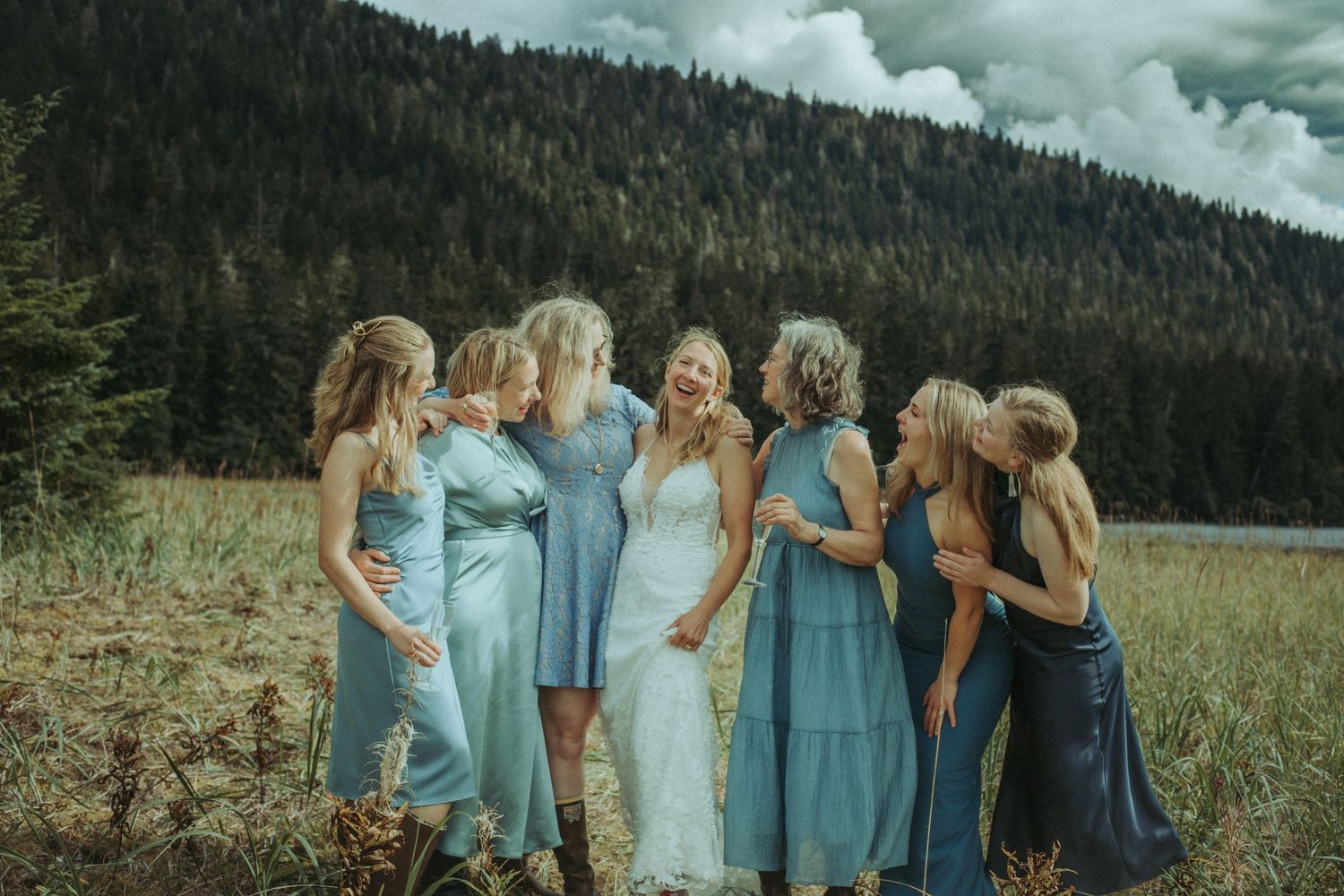 Bridesmaids in various shades of blue dresses gather closely in a beachgrass field in Alaska.