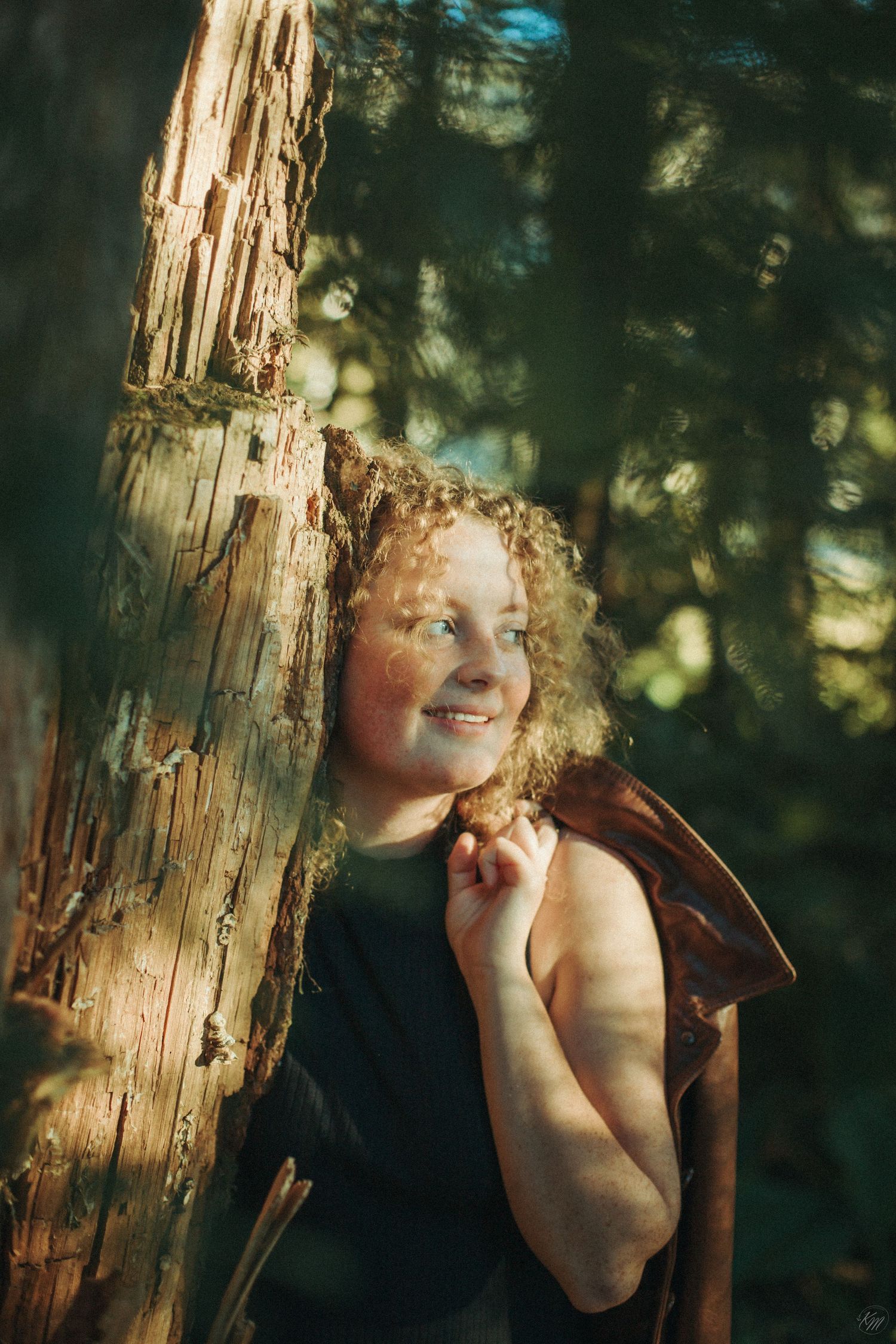Casual outdoor portrait leaning against a weathered tree trunk in dappled sunlight.