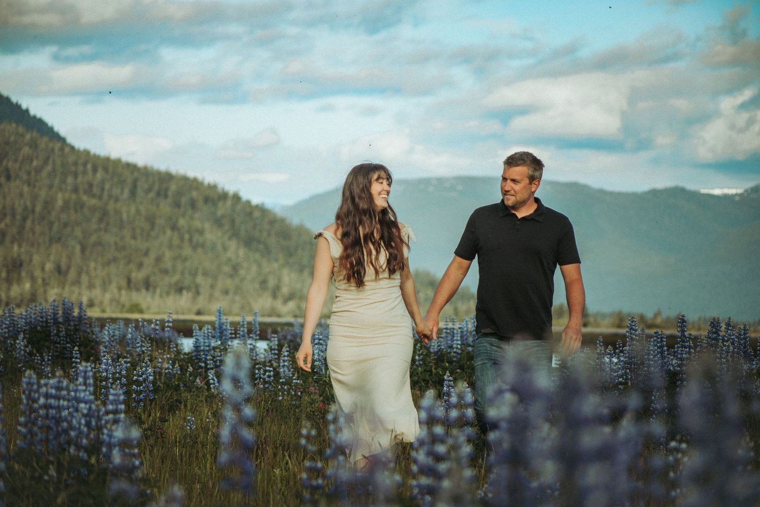 An couple holds hands while standing in a field of purple lupine with mountains and cloudy skies in the background in Southeast Alaska.
