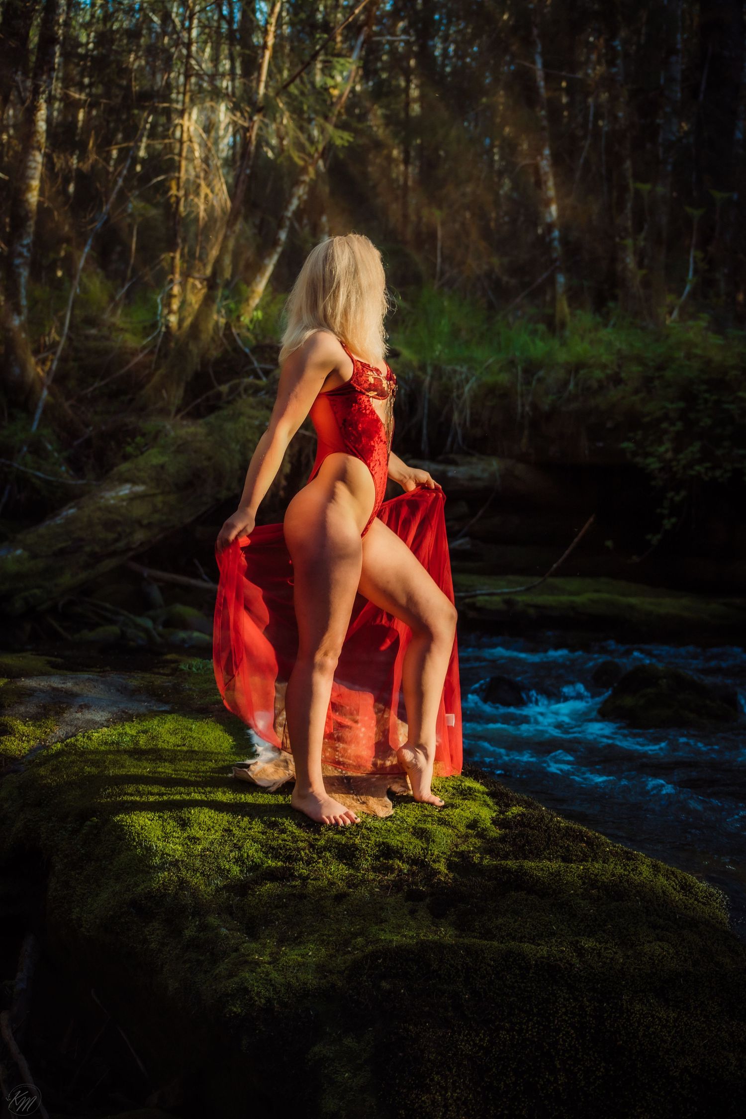 Blonde woman in red lingerie and sheer skirt standing on mossy rocks by forest stream.