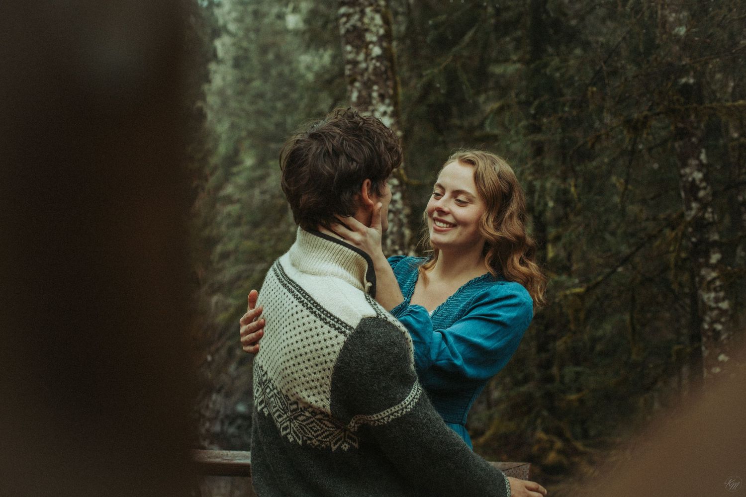A romantic couple laughs on a wooden deck surrounded by misty evergreen forest, wearing a sweater and denim blue dress in Southeast Alaska