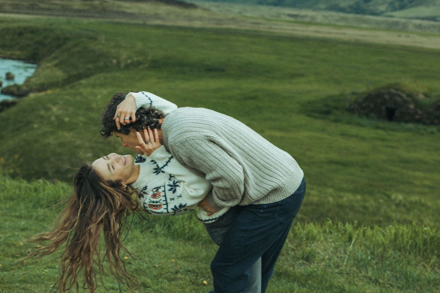Couple sharing intimate moment in green mountain landscape with dramatic lighting while wearing cozy nordic sweaters for Iceland Engagement.